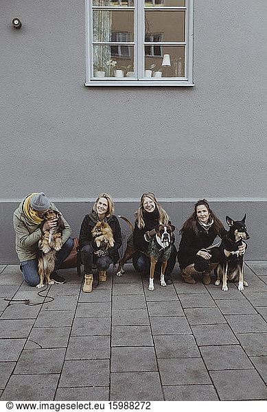 Full length portrait of smiling pet owners with dogs on footpath against wall in city
