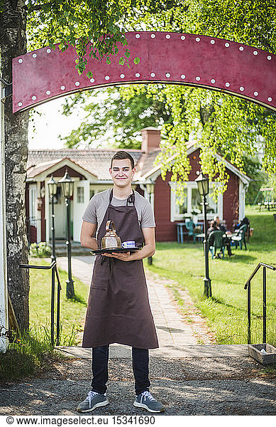 Full length portrait of smiling young waiter holding serving tray while standing at outdoor cafe entrance