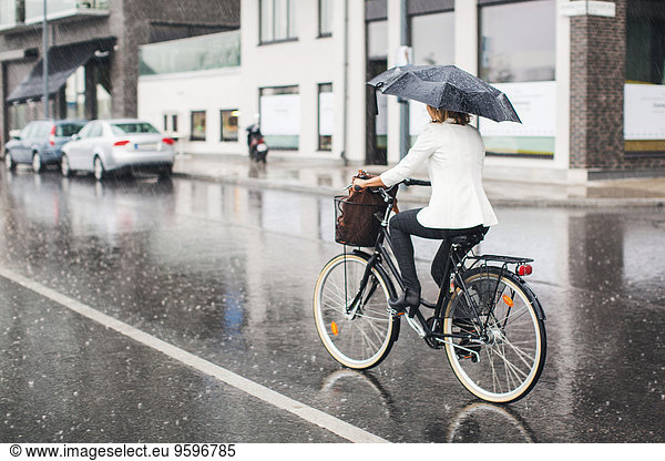 Full length rear view of businesswoman riding bicycle on wet city street during rainy season