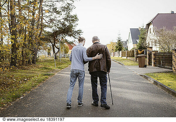 Full length rear view of caretaker supporting retired senior man walking with cane on road