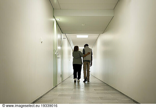 Full length rear view of female caregiver walking with arm around senior man in corridor at nursing home