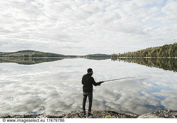Full length rear view of man fishing in lake with reflection of clouds