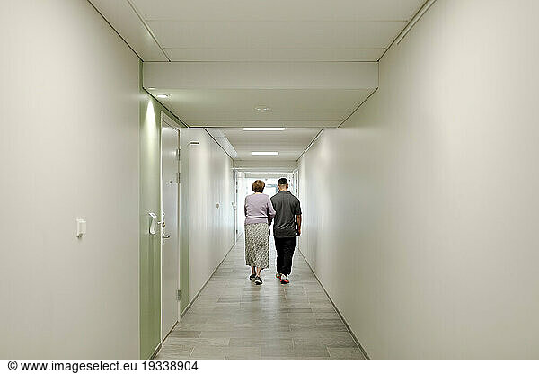 Full length rear view of senior woman walking arm in arm with male caregiver at retirement home corridor