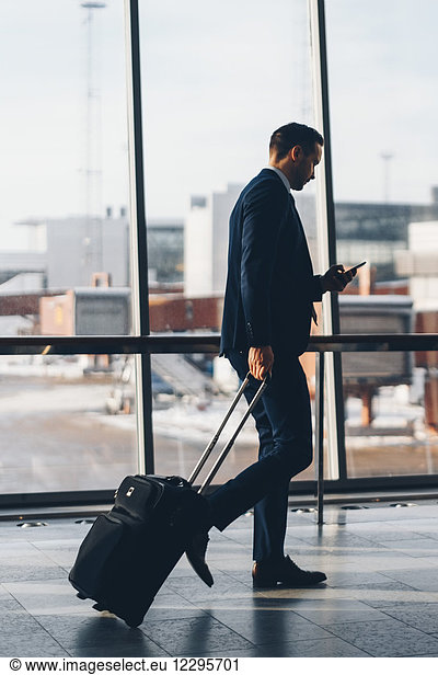 Full length side view of businessman with luggage using mobile phone while walking in airport