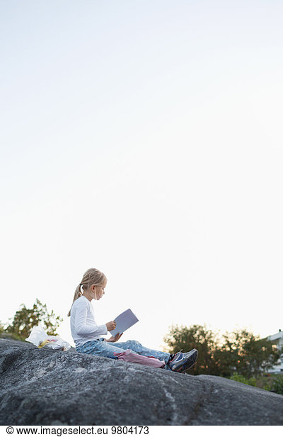 Full length side view of girl doing homework while sitting on rock against clear sky