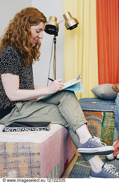 Full length side view of redhead businesswoman writing on clipboard while sitting in lobby at creative office