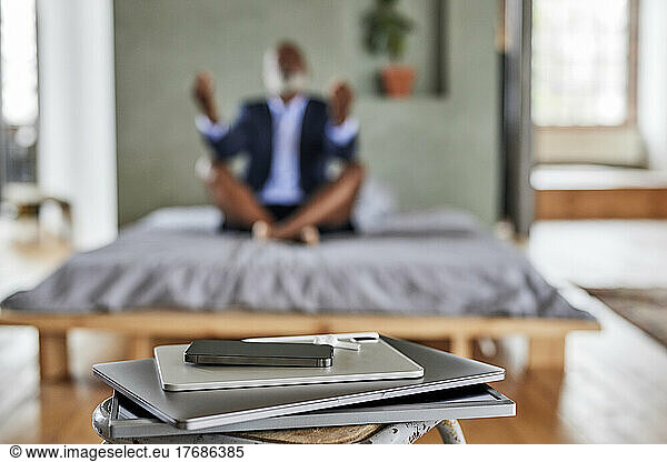 Gadgets on table with businessman meditating on bed at home