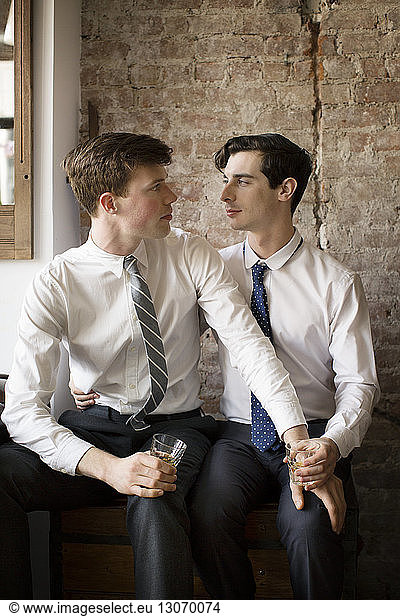 Gay couple looking at each other while sitting against brick wall at home
