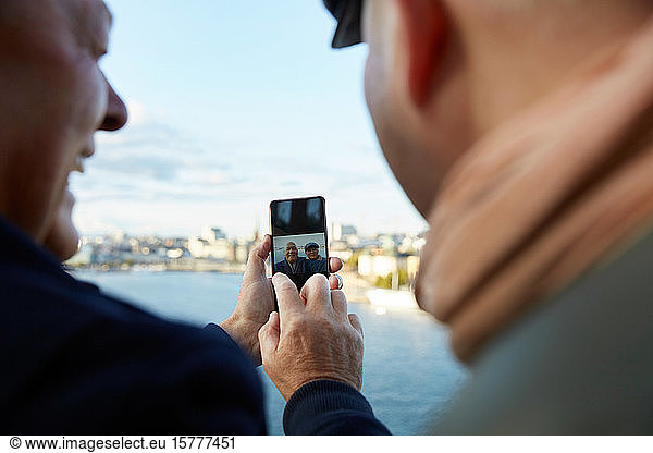 Gay couple taking selfie with mobile phone against sky in city