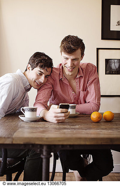 Gay couple using smart phone while sitting by table at home