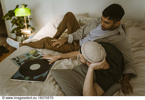Gay couple with vinyl record talking to each other while lying on bed at home