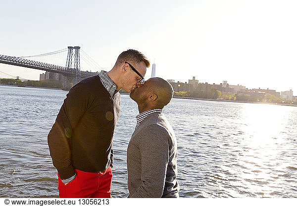 Gay men kissing while standing against East River on sunny day