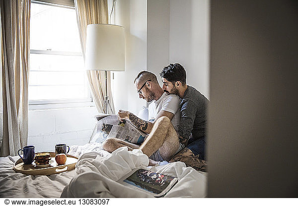 Gay men reading newspaper while sitting on bed at home
