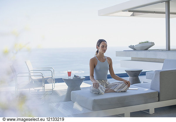 Gelassene Frau meditiert im Lotussitz auf modernem  luxuriösem Vorzeigesofa auf der Terrasse mit Meerblick