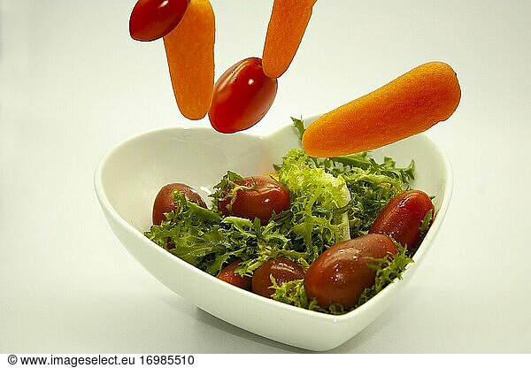 Gemüse  das in eine herzförmige Schale mit rohem veganem Bio-Salat aus Lecguga  roten Kirschtomaten und Karotten fällt.