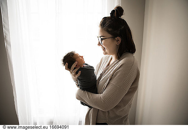 Gen Z Hipster Mom Smiles at Swaddled Newborn in Front of Window