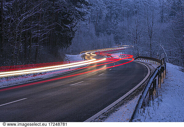 Germany  Bavaria  Spessart  Light trail on winding road through majestic winter forest at dusk
