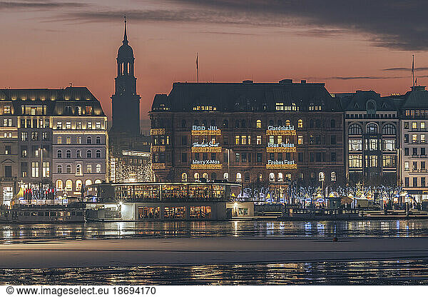 Germany  Hamburg  Christmas decorations hanging on city buildings at dusk with Alster Lake in foreground
