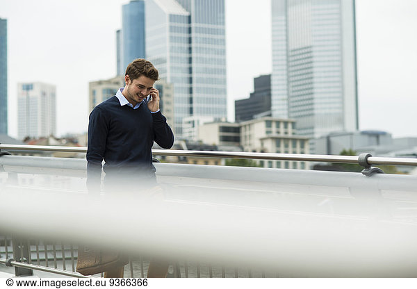 Germany  Hesse  Frankfurt  young businessman walking on a bridge telephoning with his smartphone