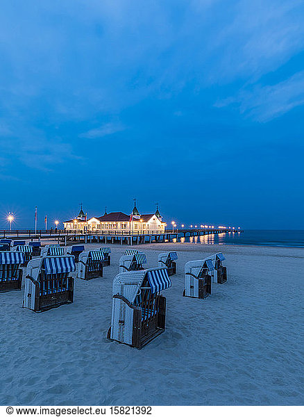 Germany  Mecklenburg-Western Pomerania  Heringsdorf  Hooded beach chairs on sandy coastal beach at dusk with illuminated pier in background