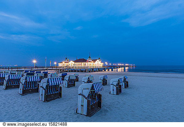 Germany  Mecklenburg-Western Pomerania  Heringsdorf  Hooded beach chairs on sandy coastal beach at dusk with illuminated pier in background