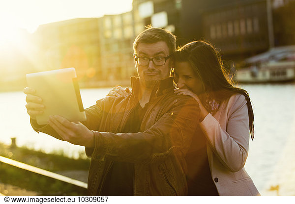 Germany  Muenster  couple taking a selfie with digital tablet at the harbour at backlight