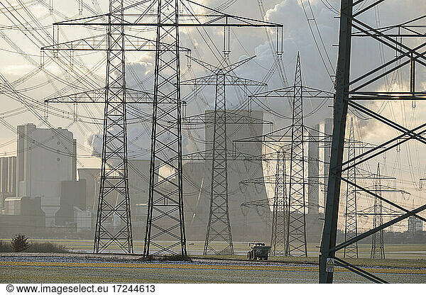Germany  North Rhine Westphalia  Niederaussem  Tractor passing by electricity pylons and lignite power plant at sunset