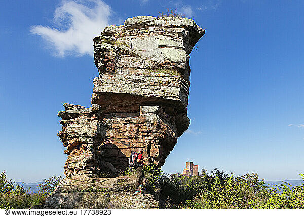 Germany  Rhineland-Palatinate  Senior hiker standing in front of sandstone rock formation in Palatinate Forest with Trifels Castle in distant background
