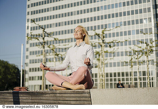Geschäftsfrau beim Meditieren im Lotussitz auf einer Bank vor einem Gebäude an einem sonnigen Tag