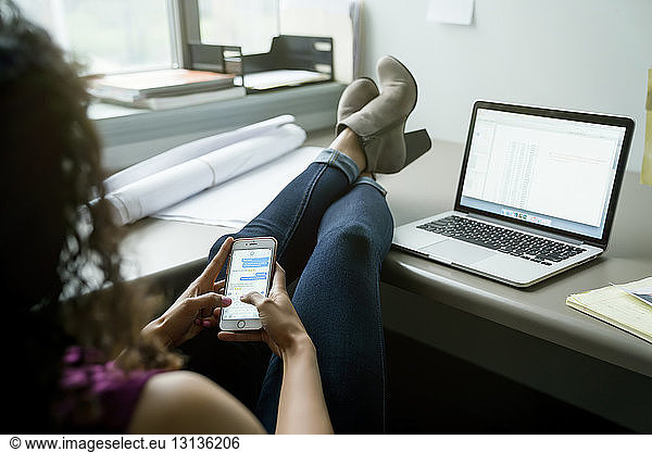 Geschäftsfrau benutzt Mobiltelefon  während sie sich am Schreibtisch im Büro entspannt