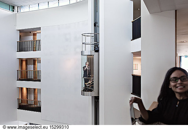 Geschäftsfrau steht auf dem Balkon  während ein Geschäftsmann mit seinem Sohn im Aufzug im Büro steht