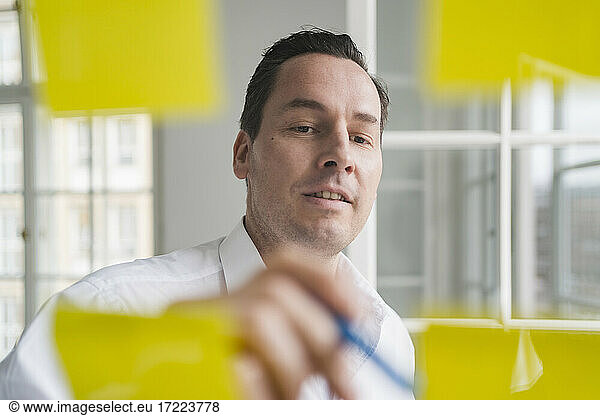 Geschäftsmann beim Brainstorming mit Notizen auf transparenter Wischtafel am Arbeitsplatz