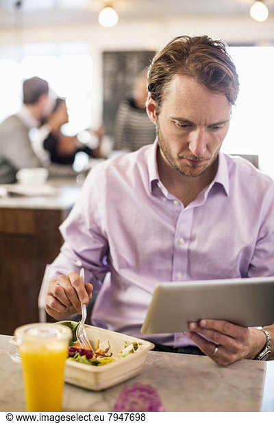 Geschäftsmann  der beim Frühstück mit Kollegen im Hintergrund im Restaurant das digitale Tablett betrachtet