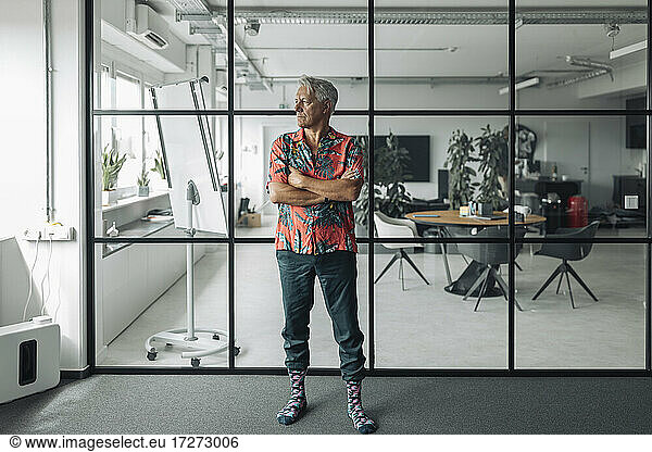 Geschäftsmann in Freizeitkleidung steht mit verschränkten Armen vor einer Glaswand im Büro