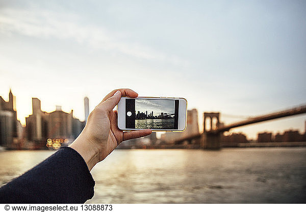 Geschnittene Hand einer Frau  die in der Dämmerung die Skyline und die Brooklyn Bridge fotografiert