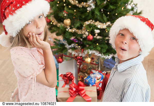 Girl and boy wearing santa hats in front of christmas tree looking at camera excitedly