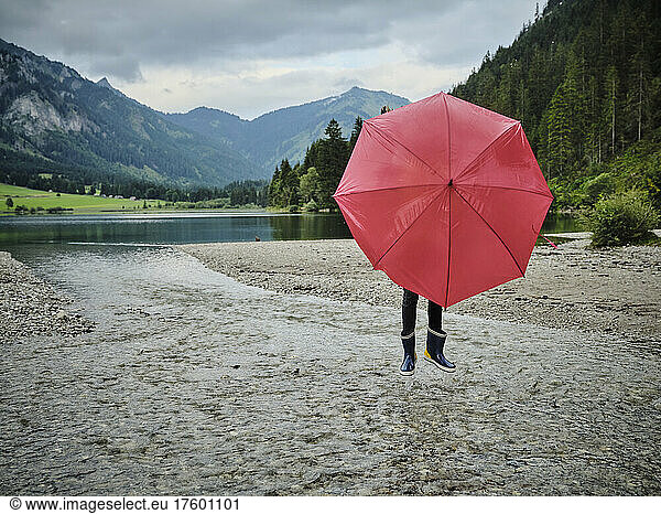 Girl covering face behind red umbrella standing on water at lakeshore