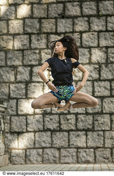 Girl dancing with ballet shoes and in a pose mid-air against a stone wall at a public park; Hong Kong  China