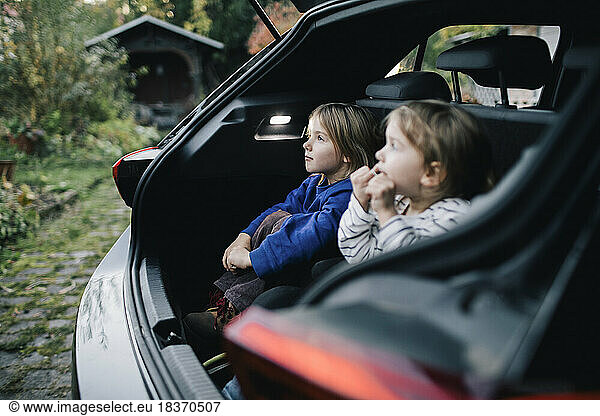 Girl day dreaming by sister while sitting in electric car trunk