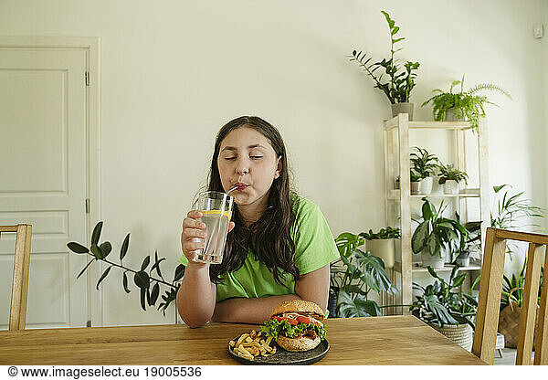 Girl drinking soda from glass with burger on table at home