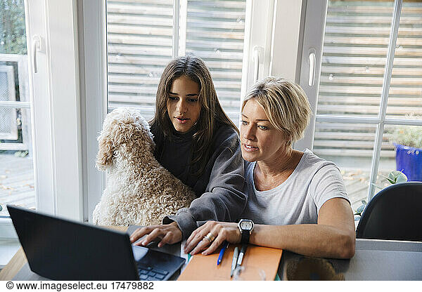 Girl e-learning on laptop while sitting with mother and dog at home