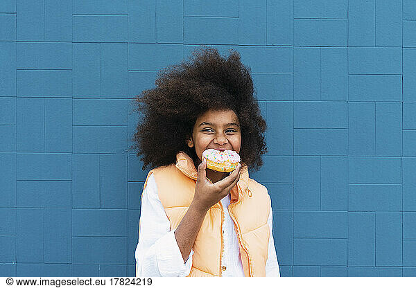 Girl eating doughnut standing in front of blue wall