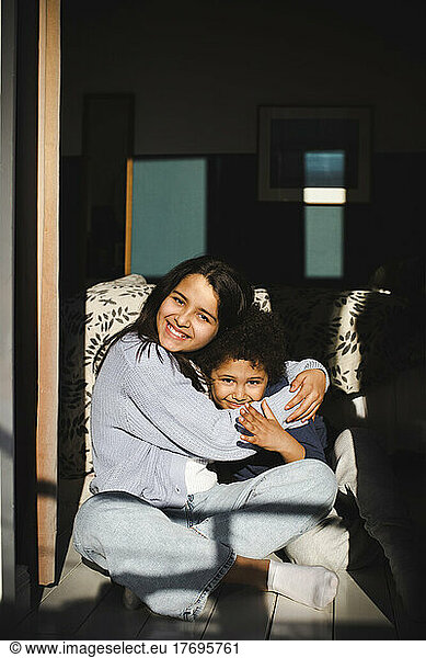 Girl embracing brother while sitting cross-legged in sunlight at home