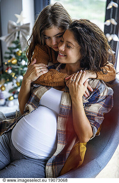 Girl embracing expectant mother sitting on chair at home
