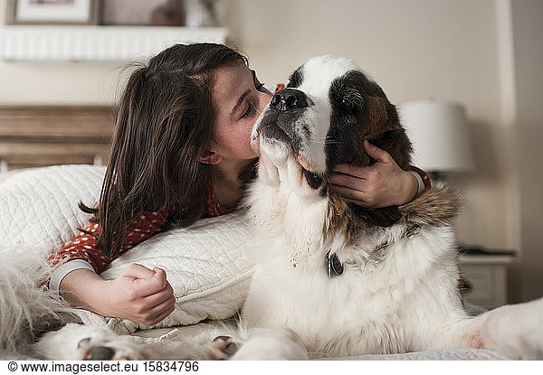Girl gives dog big kiss on the cheek while laying on bed at home