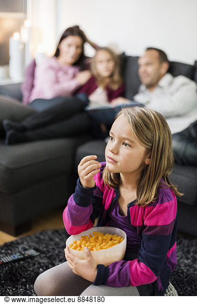 Girl having snacks while watching TV on floor with family in background