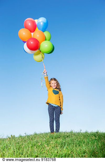 Girl holding bunch of balloons on grassy hill
