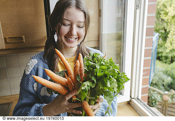 Girl holding bunch of carrots near window at home
