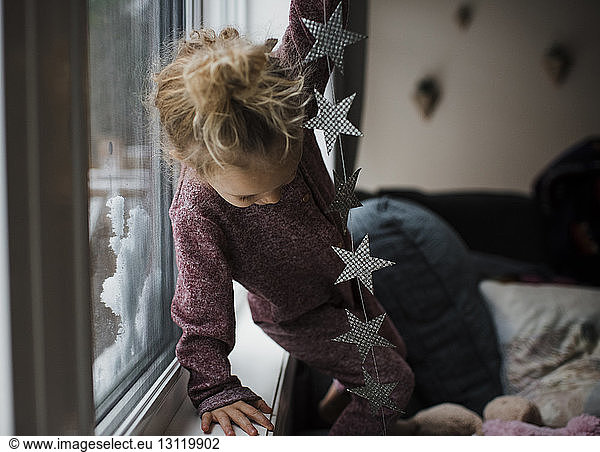 Girl holding star shape decoration while standing on sofa by window in living room