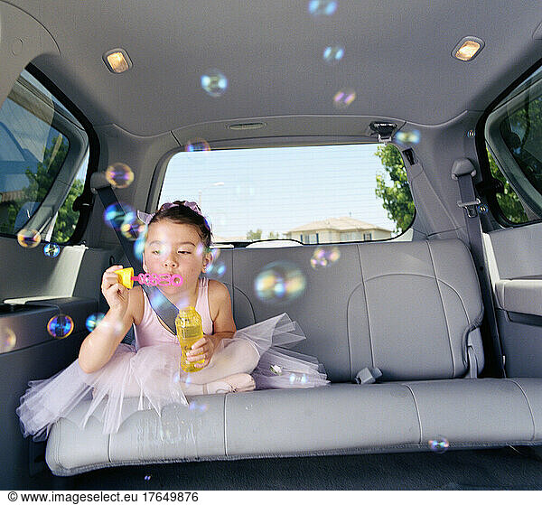 Girl (6-7) in ballet outfit blowing bubbles in car